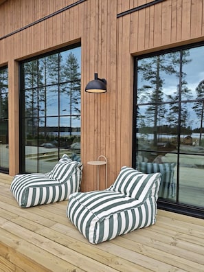 Terraza de madera con dos sillones orejeros a rayas y una mesita, con vista a un cuerpo de agua rodeado de árboles.