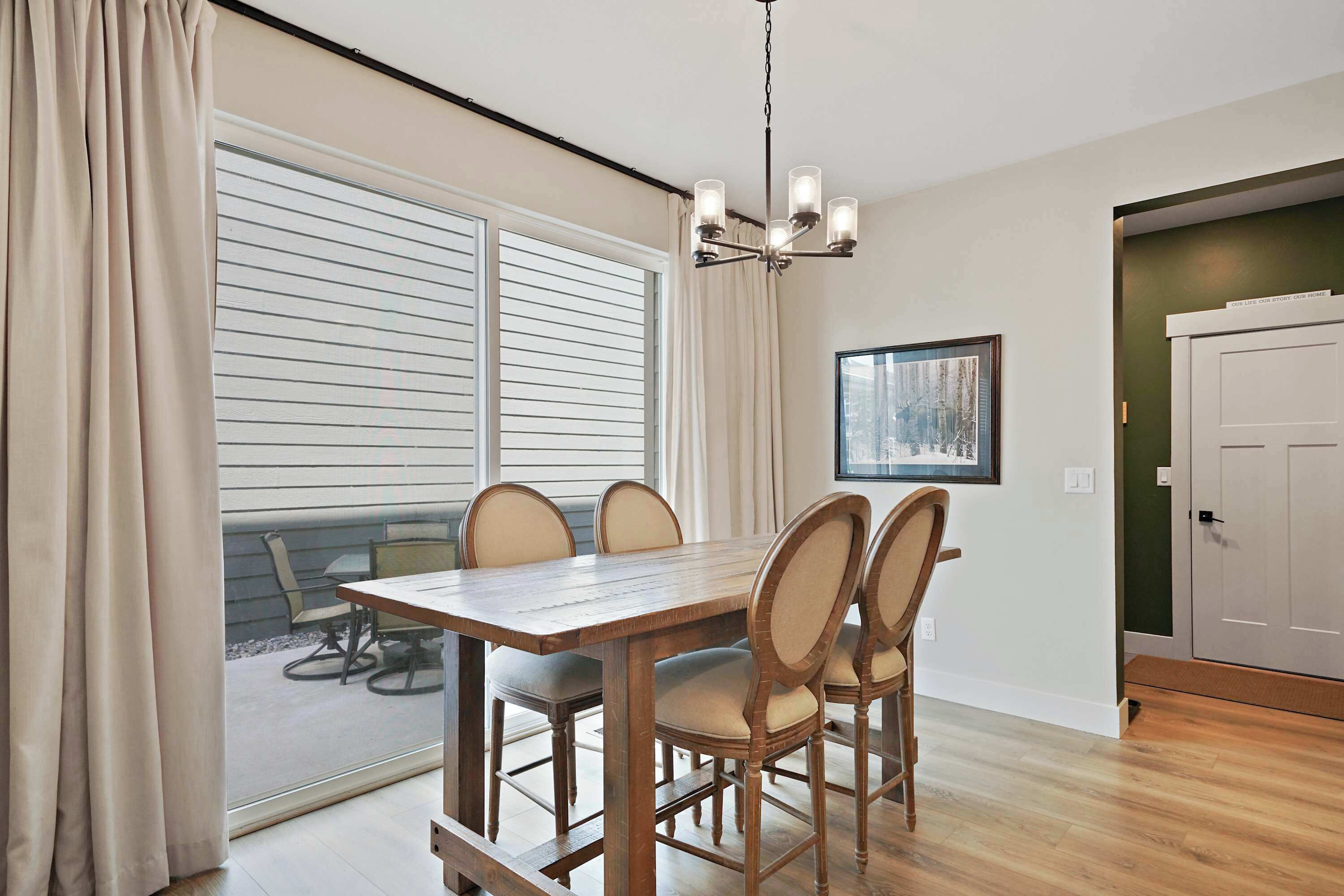 A dining area with a wooden table and chairs, a chandelier, a window with blinds, and a door.