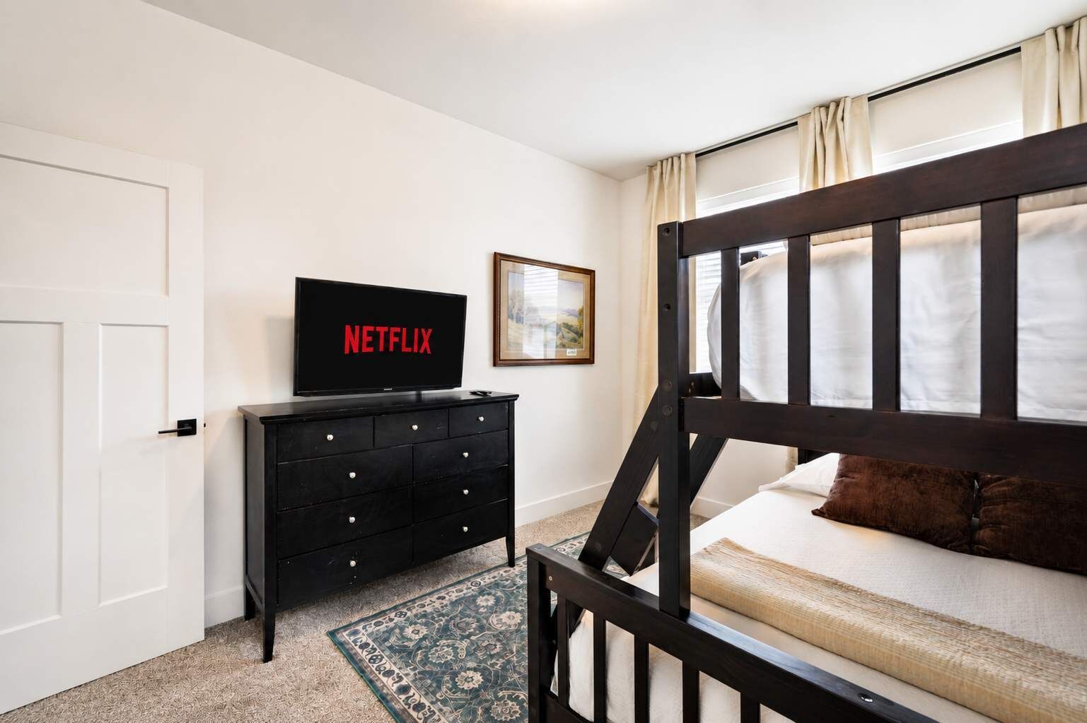 Un dormitorio con cama, televisor sobre una cómoda, puerta y ventana con cortinas.