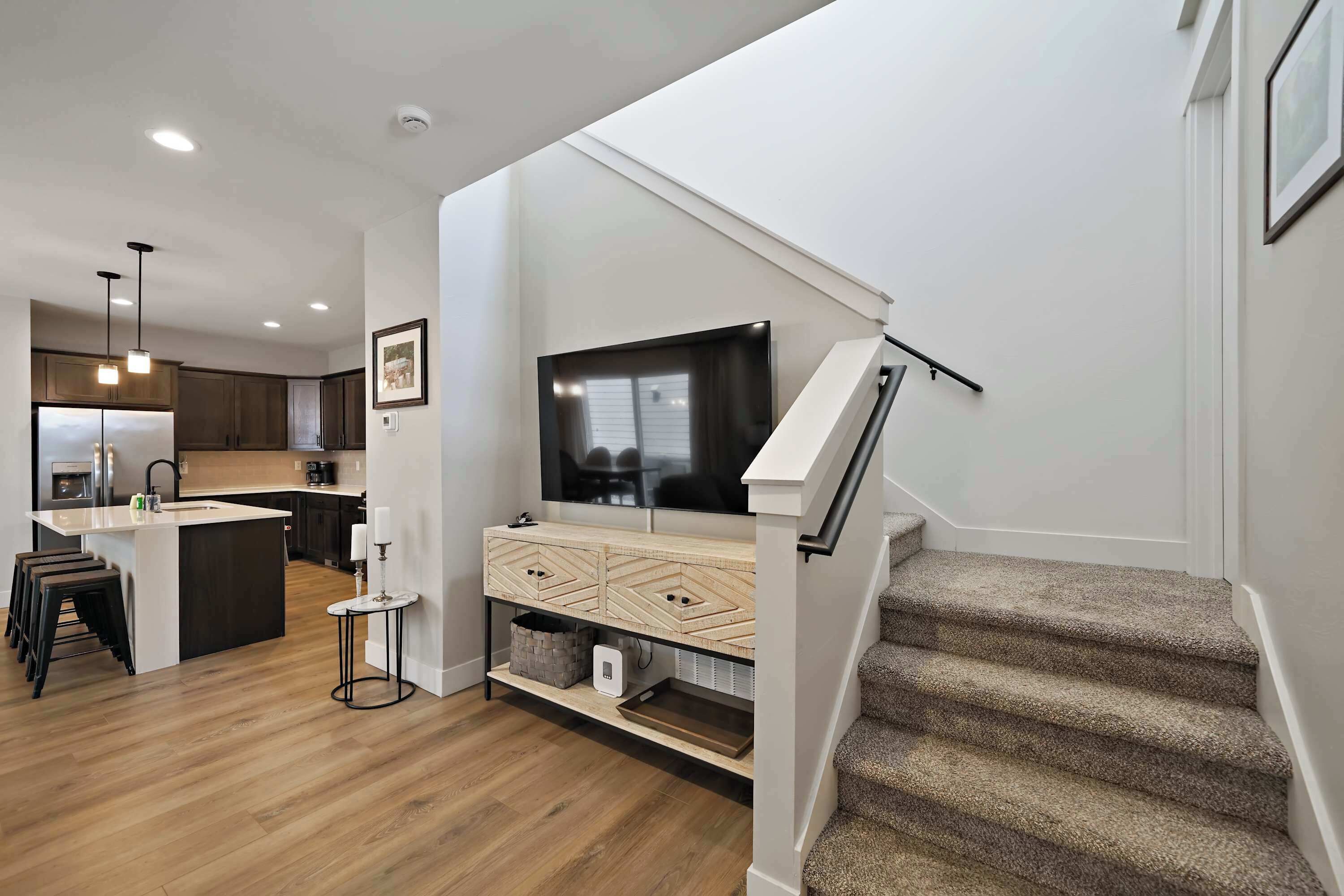 Una cocina moderna con una isla central, un comedor con taburetes, una escalera con barandilla y un televisor de pantalla plana montado en la pared.