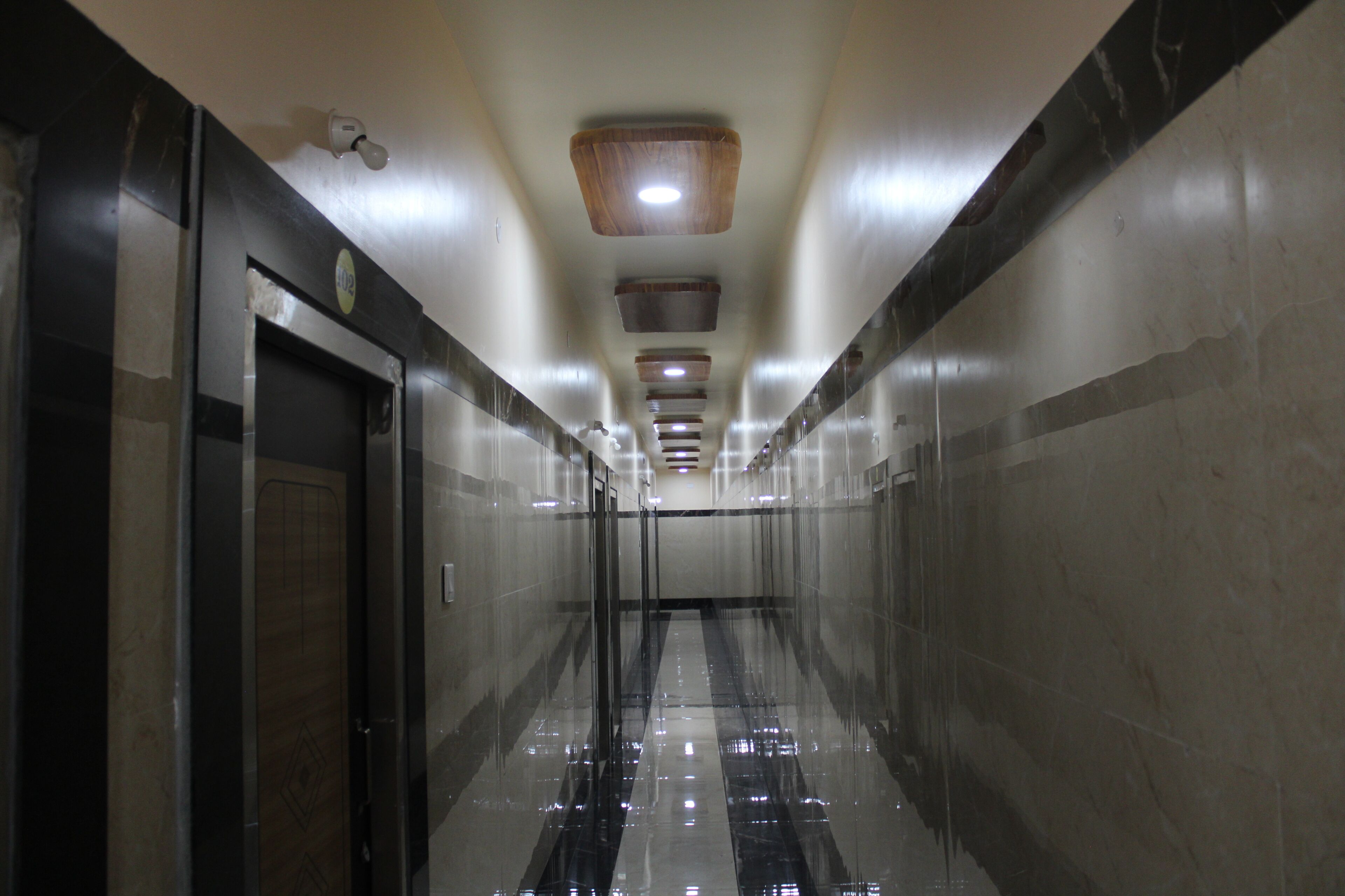 A corridor with a reflective floor, a door on the left, and a series of lights on the ceiling.