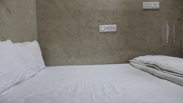 A single bed with white bedding in a room with a marble wall and a power outlet. - Srivari Lodge (Aarkadu)