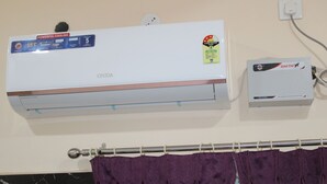 A wall-mounted air conditioner with a power savings guide sticker, a wall socket, and a curtain rod with purple curtains. - Srivari Lodge (Aarkadu)