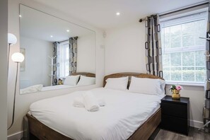 A hotel room with a large bed, a bedside table, a nightstand, a mirror, and a window with blinds. - Spacious 5-bed Home in Milton Keynes by Renzo, Driveway Parking, Close to M1 (Milton Keynes)