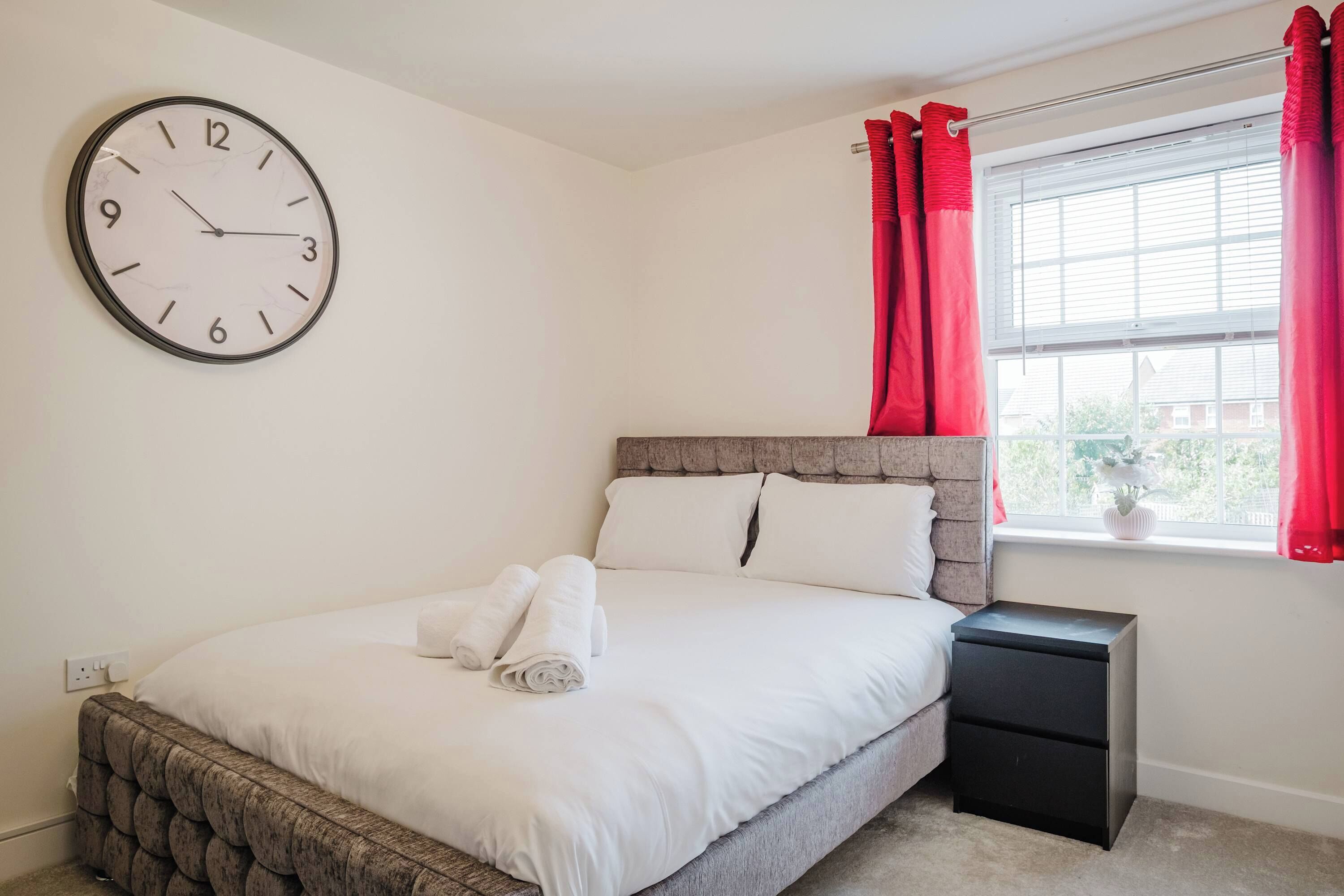 Een slaapkamer met een bed, nachtkastje, een klok aan de muur en een raam met gordijnen.