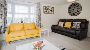 Un salon avec un canapé jaune, un canapé en cuir noir, une table basse blanche et une grande horloge murale.