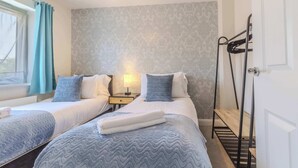 Ein Hotelzimmer mit zwei Betten, einem Nachttisch, einer Lampe, einem Fenster mit Vorhängen und einer Tür mit Handtuchhalter.