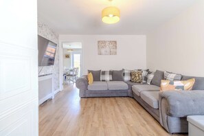 Een woonkamer met een grijze hoekbank, een flatscreen televisie aan de muur en een houten vloer.