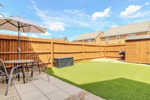 Een achtertuin met kunstgras, een houten omheining, een barbecue en tuinmeubilair.