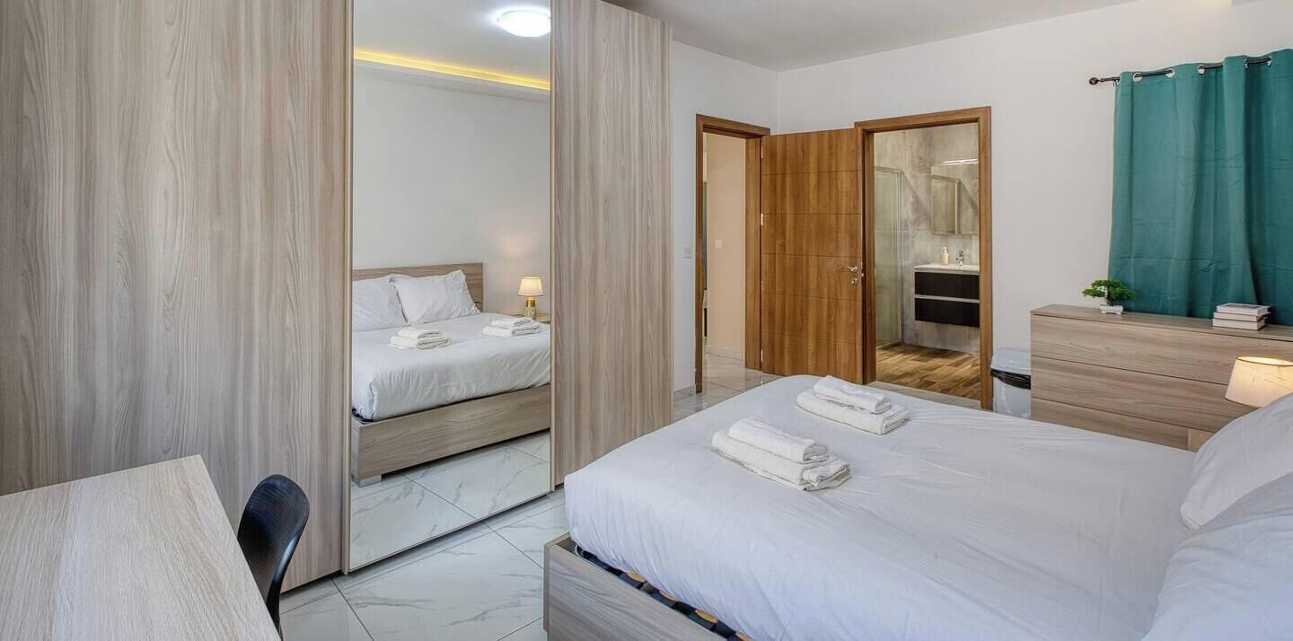 Ett hotellrum med en säng, ett skrivbord, en stol, en garderob och ett badrum med en spegel och ett handfat.