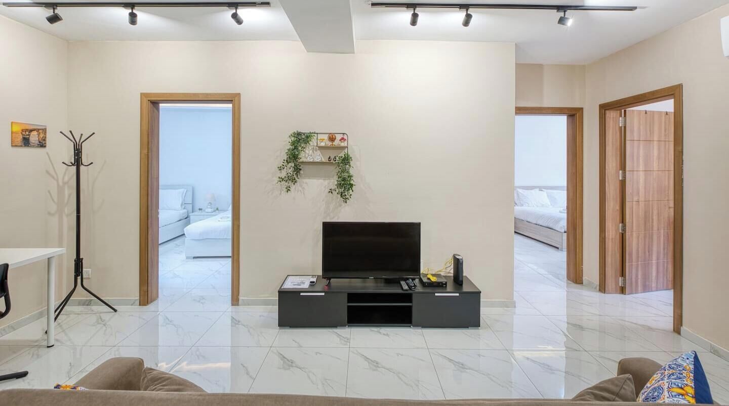 Een moderne woonkamer met een flatscreen televisie, een salontafel en een bank. Er staat een kapstok en een plant aan de muur.