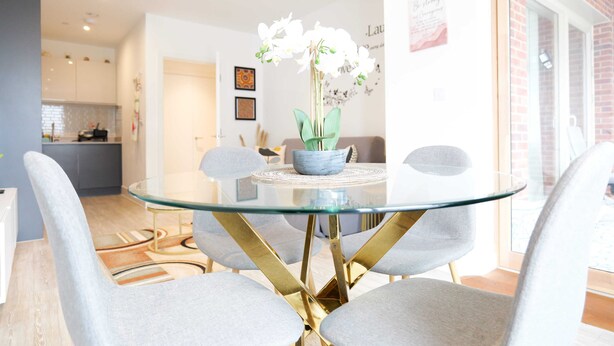 Un comedor moderno con una mesa de vidrio, sillas tapizadas de color gris y un florero con flores blancas.