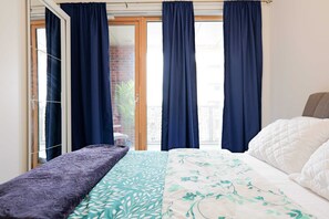 Quarto com cama, um banco e porta de vidro deslizante que dá para uma varanda.