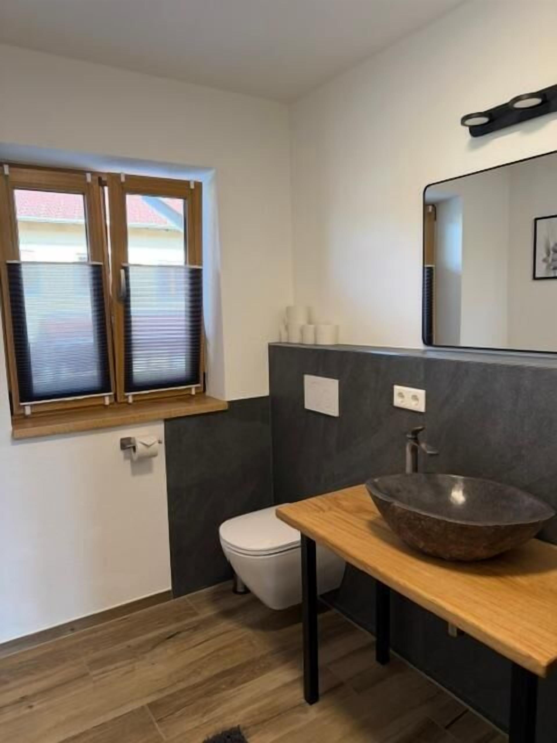 A bathroom with a wooden vanity, a vessel sink, a toilet, and a window with blinds.