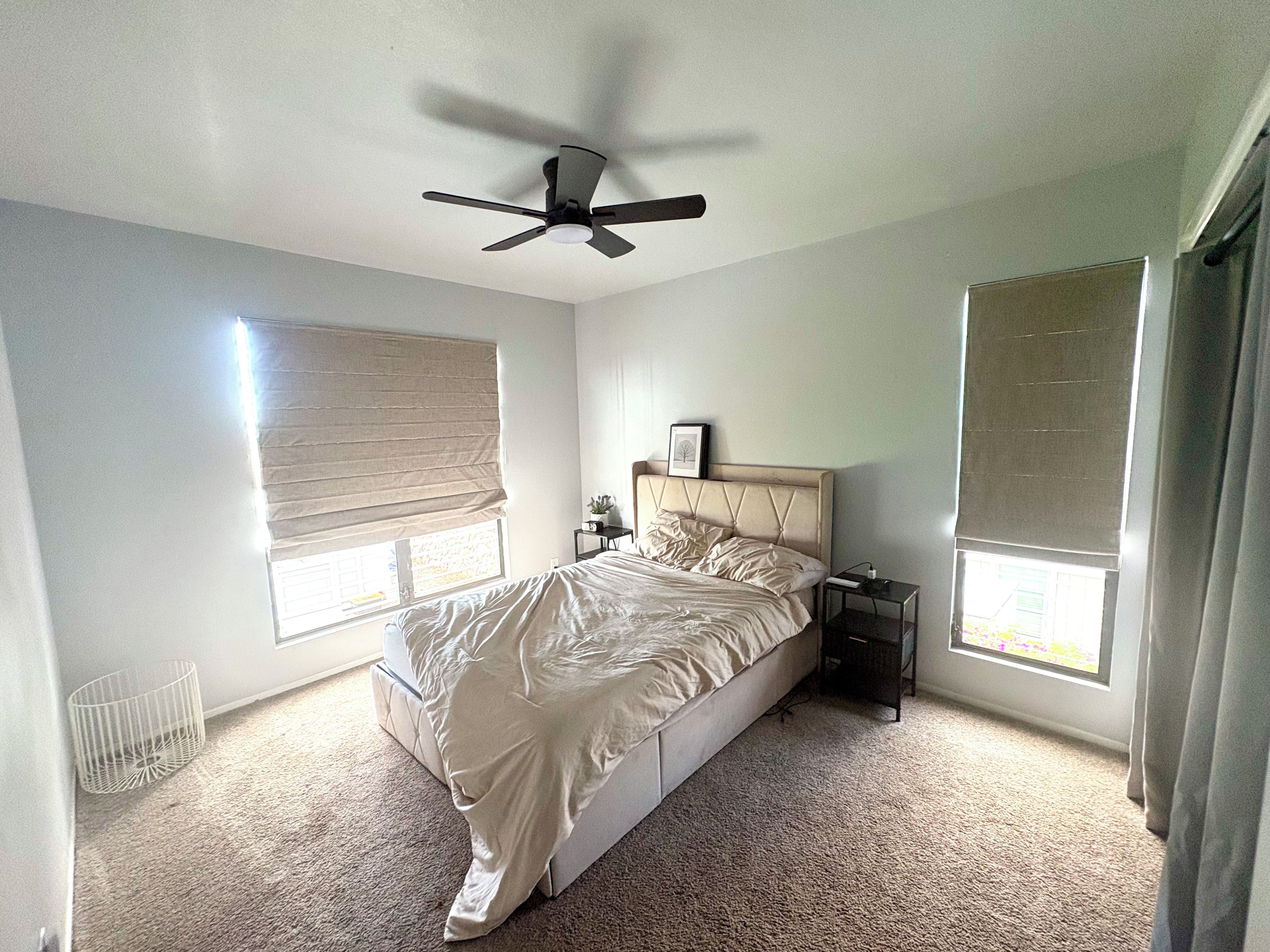 A bedroom with a bed, a ceiling fan, a nightstand, a window with blinds, and a radiator.
