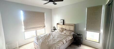 A bedroom with a bed, a ceiling fan, a nightstand, a window with blinds, and a radiator.