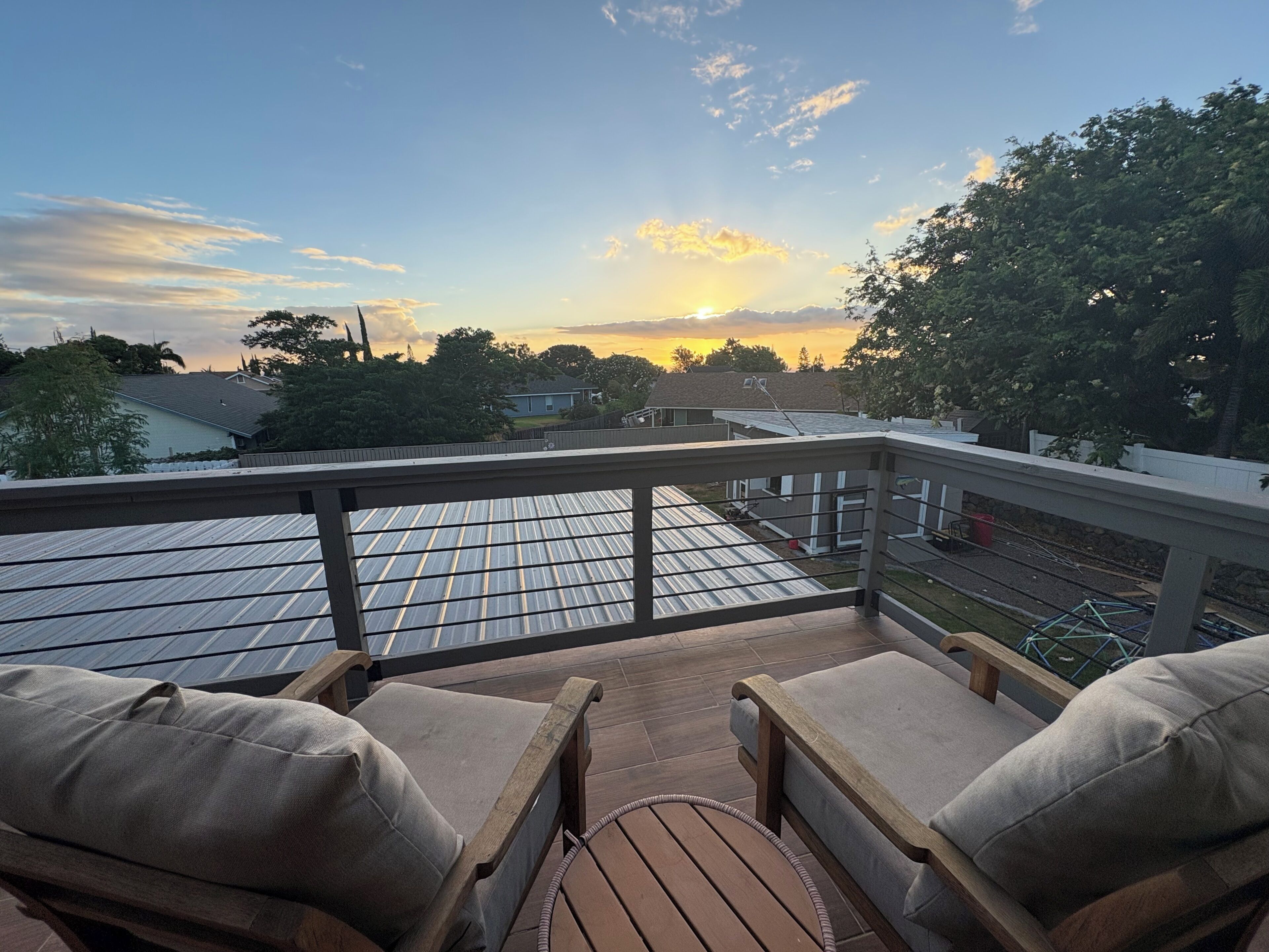 A balcony with two cushioned chairs, a wooden deck, and a view of a residential area during sunset.