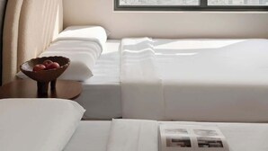 Habitación de hotel moderna con vista a la ciudad, una ventana grande con persianas, una cama, una mesita de noche con un frutero y una obra de arte colgada en la pared.