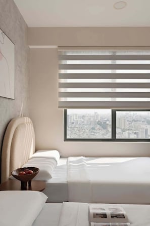 A modern hotel room with a city view, a large window with blinds, a bed, a bedside table with a bowl of fruit, and a wall-mounted artwork. - Corner 2BR/3Beds • River View • Walk to Bui Vien (Thành phố Hồ Chí Minh)