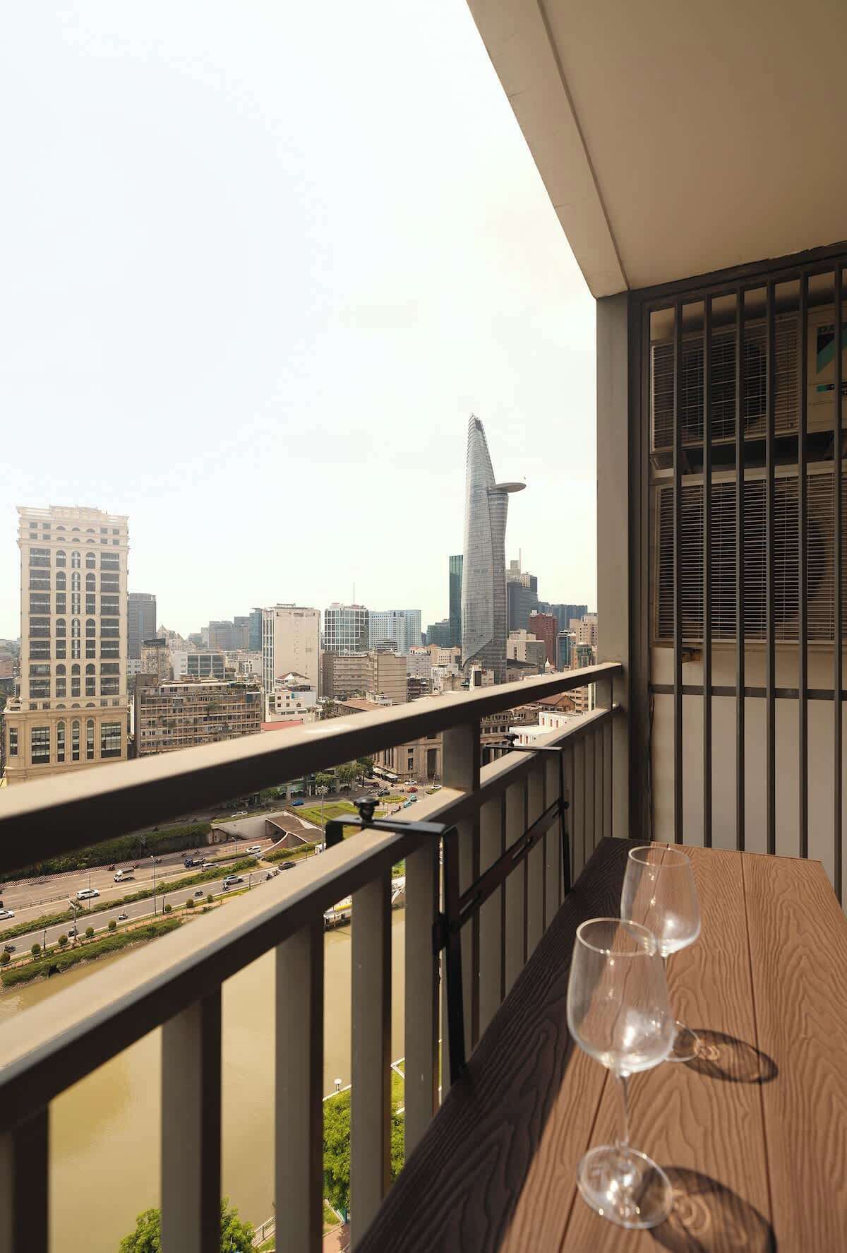 Ein Balkon mit Blick auf eine Stadtlandschaft, darunter ein markanter Wolkenkratzer, und zwei leere Weingläser auf dem Tisch.