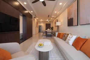 A modern living room with a sofa, a coffee table with fruit, a TV, and a dining area with chairs and a table. - 3BR+2BA | 4 Beds | Sleeps 11 | Bitexco View | D1 (Thành phố Hồ Chí Minh)