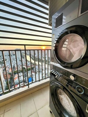 A laundry area with stacked washers and dryers, a balcony with a view of a cityscape, and a window with horizontal blinds. - Stay Together | 5BRs/4BA on Same Floor for Groups (Thành phố Hồ Chí Minh)