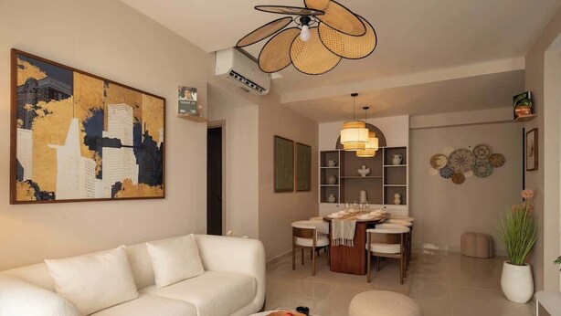 Ein modernes Wohnzimmer mit einer Couch, einem runden Couchtisch mit Früchten, einem Essbereich mit Tisch und Stühlen sowie einem Deckenventilator.