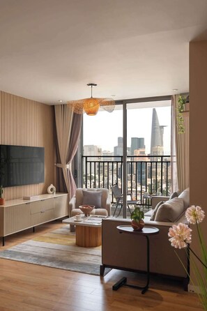 Ein modernes Wohnzimmer mit großem Fernseher, bequemen Sitzmöglichkeiten, einem Couchtisch und Blick auf eine Stadtkulisse durch große Fenster.