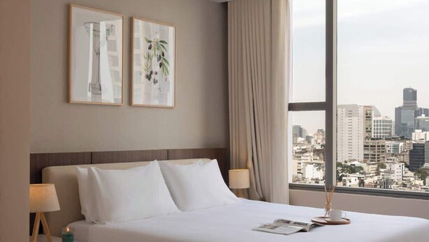 Una camera d'albergo moderna con un letto grande, un comodino, una lampada, un tavolino con una tazza e un libro, e vista sulla città attraverso ampie finestre.