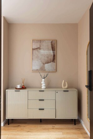 A room with a beige wall, a wooden floor, a white cabinet with drawers, a framed abstract artwork, and a vase with branches. - Large 3BRs+2WCs | City & D1 View | 5 min BuiVien (Thành phố Hồ Chí Minh)