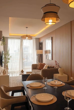 A modern living room with a dining area, featuring a sofa set, a dining table with plates and glasses, and a large window with sheer curtains. - 4BR · 5 Beds · 2BA · Fireworks/Bitexco View · D4 (Thành phố Hồ Chí Minh)