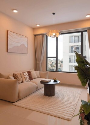 Ein modernes Wohnzimmer mit einer Couch, einem runden Couchtisch, einem großen Fenster mit Blick auf Gebäude und einer Pflanze.