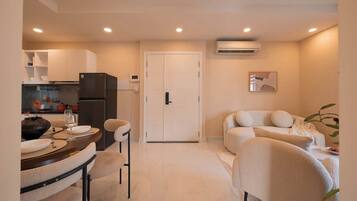 A modern living room with a sofa and chairs, a dining area with a table and chairs, a kitchen with a refrigerator, and a closed door.