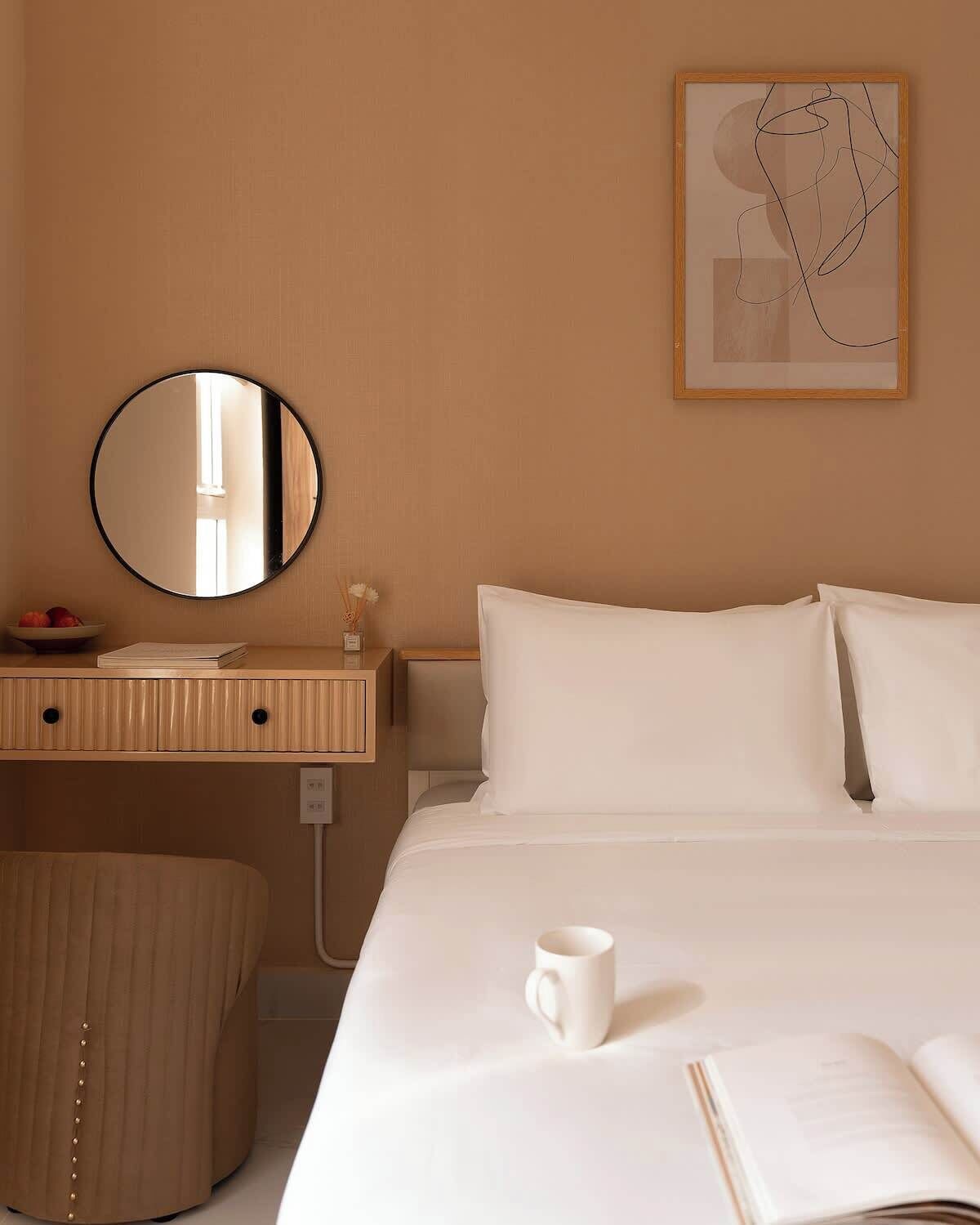 A hotel room with a bed, a round mirror, a bedside table with a book and a cup, and a wall art piece.