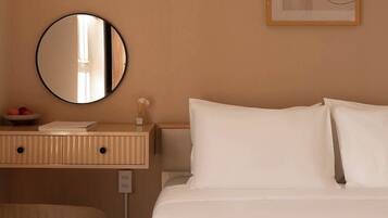 A hotel room with a bed, a round mirror, a bedside table with a book and a cup, and a wall art piece.