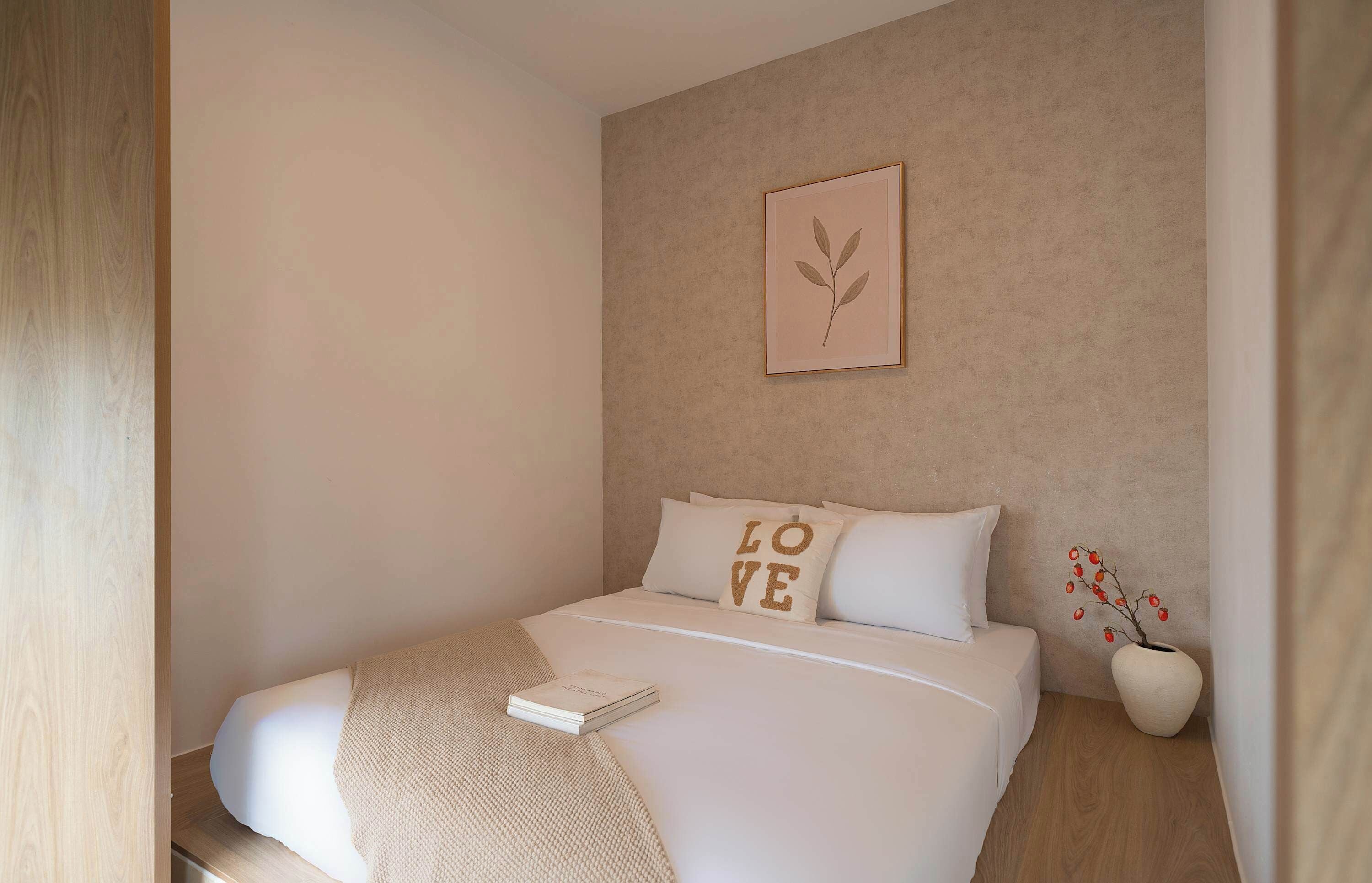 Een net opgemaakt bed met een kussen waarop 'LOVE' staat, een klein plantje in een lijstje aan de muur, en een boek op het bed.