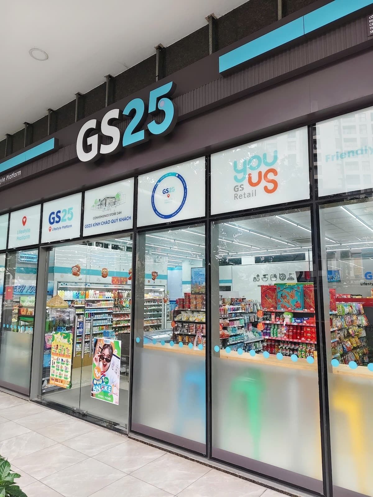 Un magasin GS25, de type épicerie de proximité, avec une façade en verre transparent, différents présentoirs de produits et des affiches promotionnelles.