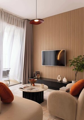 A modern living room with a flat-screen TV mounted on a wall, a round coffee table, two sofas with orange cushions, a potted plant, and a large window with sheer curtains. - 3BR | 2BA • Near D1 • River View • Dryer • HighFLR (Thành phố Hồ Chí Minh)
