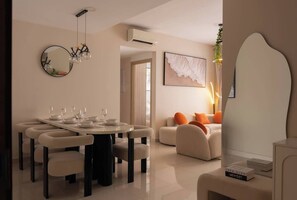 A modern living room with a dining area, featuring a round table, chairs, a sofa with orange cushions, a large abstract painting, and a decorative mirror. - 3BR | 2BA • Near D1 • River View • Dryer • HighFLR (Thành phố Hồ Chí Minh)
