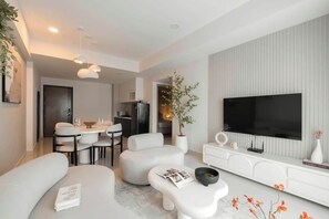 Ein modernes Wohnzimmer mit Flachbildfernseher, weißen Sofas, einer Essecke und einer dahinterliegenden Küche.