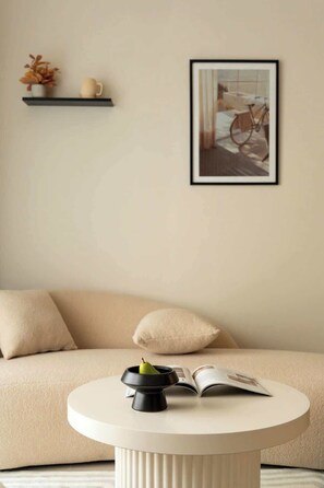 A minimalist living room with a sofa, a round table with a book and a fruit bowl, a framed picture on the wall, and a shelf with decorative items. - 2BR + 3 Beds | 1BA • Central D1 • Side River View (Thành phố Hồ Chí Minh)
