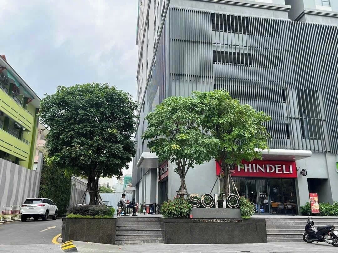 Ein modernes Gebäude mit einem großen Baum davor, einem Schild für 'SOHO' und 'PHINDELI' im Erdgeschoss und einem geparkten Auto.