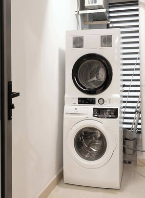 A stacked washer and dryer unit in a laundry room with a door and a window with blinds. - Luxury 3BR 2BA | Central | 2 min BuiVien | D1 (Thành phố Hồ Chí Minh)