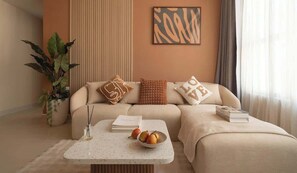 A modern living room with a sofa, a coffee table with fruits and books, a large plant, and a wall with abstract art. - Luxury 3BR 2BA | Central | 2 min BuiVien | D1 (Thành phố Hồ Chí Minh)