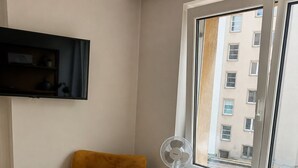 Ett rum med en senapsgul fåtölj, en väggmonterad tv, ett fönster med utsikt över byggnader, ett element och en golvlampa.