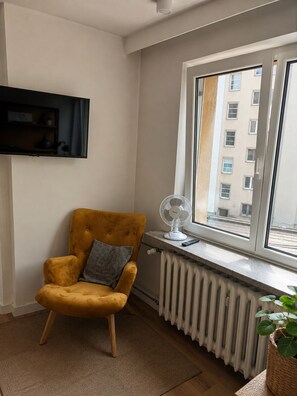 A room with a mustard-colored armchair, a wall-mounted TV, a window with a view of buildings, a radiator, and a floor lamp. - Native Apartments Starowiślna 25 (Kraków)