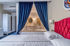 A hotel room with a large bed, a red headboard, a glass-enclosed bathroom, and blue curtains. - Czeczotka Palace Apartments Old Town (Kraków)