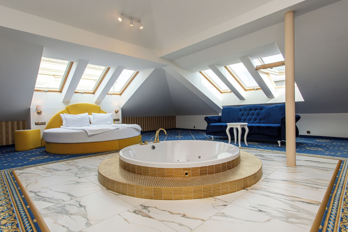 A modern bedroom with a large circular bathtub, a sofa, and a bed with a yellow headboard.