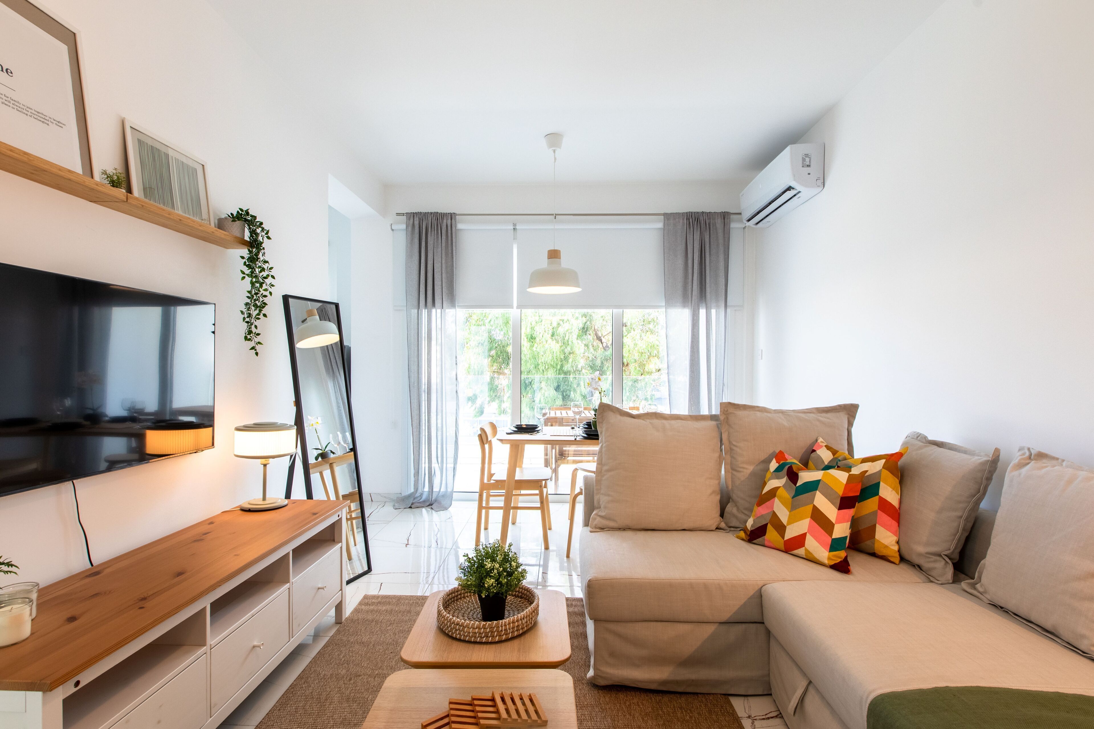 A modern living room with a flat-screen TV, a wooden side table, a sofa with patterned cushions, and a dining area with a table and chairs.