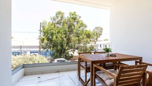 A balcony with a wooden table and chairs, a potted plant, and a view of a tree and buildings. - Platinum Breeze - 1-BR Apt. in Larnaca (Larnaka)
